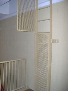 A beige wall-mounted safety ladder with a mesh guard and an open safety cage panel, positioned near a metal railing.