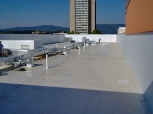 A flat rooftop with white surface and HVAC ductwork, featuring a tall building and distant mountains under a blue sky.