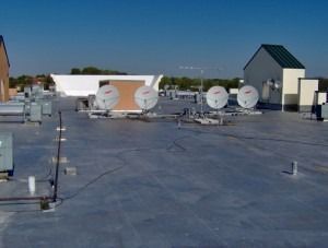 A flat gray commercial rooftop featuring four satellite dishes and building utility structures under a clear blue sky.