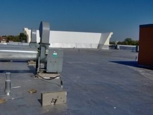 A rooftop view featuring a large grey HVAC fan unit on a metallic surface, with a white wall in the background.