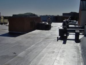 A flat commercial roof with HVAC equipment, a brick partition, and utility pipes under a clear blue sky.