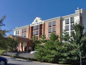 A low-angle view of a multi-story, beige brick Hyatt Place hotel under a clear blue sky, surrounded by green trees.