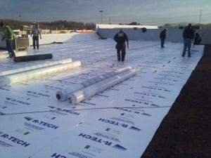 Workers install white R-Tech foam insulation panels on a large, flat rooftop under a clear sky.
