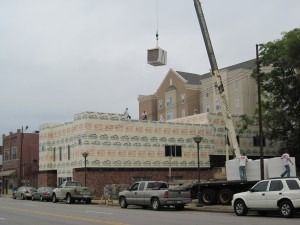 A crane lifts a HVAC unit onto the roof of a building under construction, wrapped in patterned building paper.