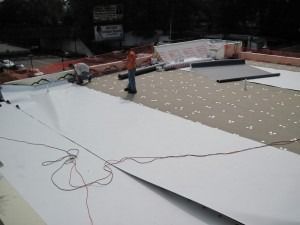A worker installs a white TPO roofing membrane over insulation boards on a flat commercial roof.
