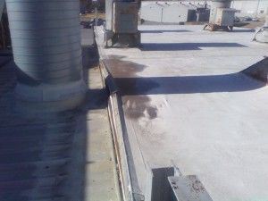 A flat, light-colored industrial roof showing a metal partition, a large cylindrical vent, and a puddle of standing water.