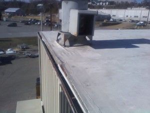An outdoor view from above showing a flat white commercial roof, a metal vent, and a damaged edge along the building wall.
