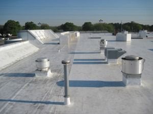A flat white commercial roof with several ventilation units and pipes under a clear blue sky.
