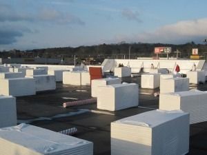 Stacked white insulation boards sit on a flat, dark roof under a bright blue sky with a distant sign.