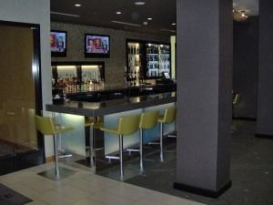 A modern hotel bar with sleek countertops, backlit shelving, and several yellow bar stools in a dimly lit room.