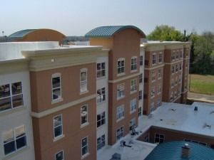 A multi-story tan apartment building with cream trim and arched green roof accents under a clear blue sky.