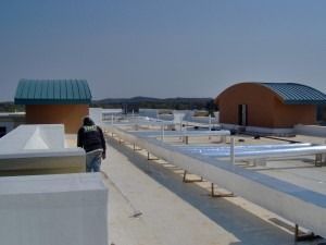 A worker walks across a flat building roof featuring long white ducts and two small structures with curved blue roofs.