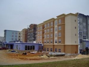 A multi-story hotel building under construction, featuring tan and blue sections, unfinished siding, and a dirt yard.
