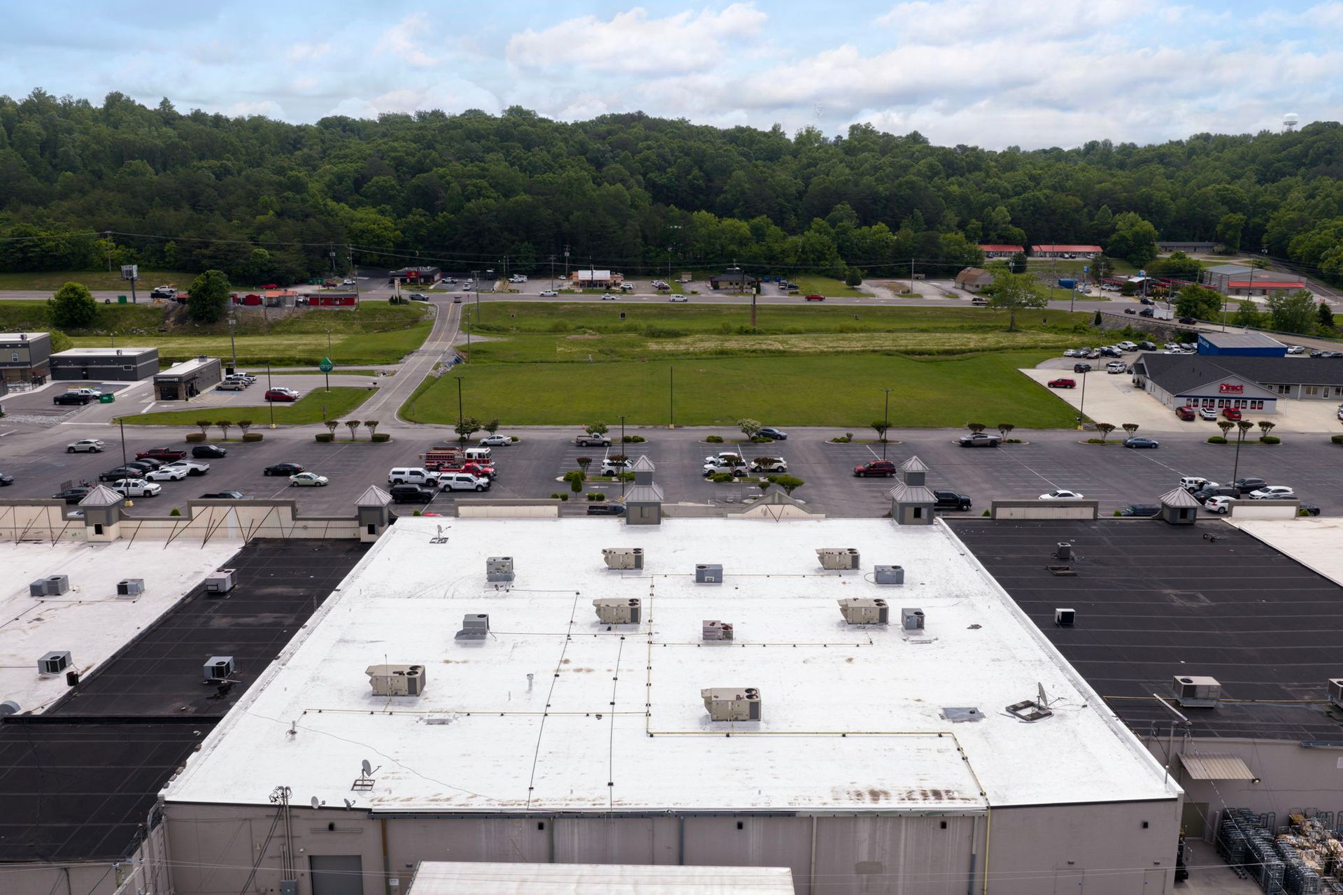 Aerial view of a large parking lot beside a white-roofed building and grassy field with trees beyond.