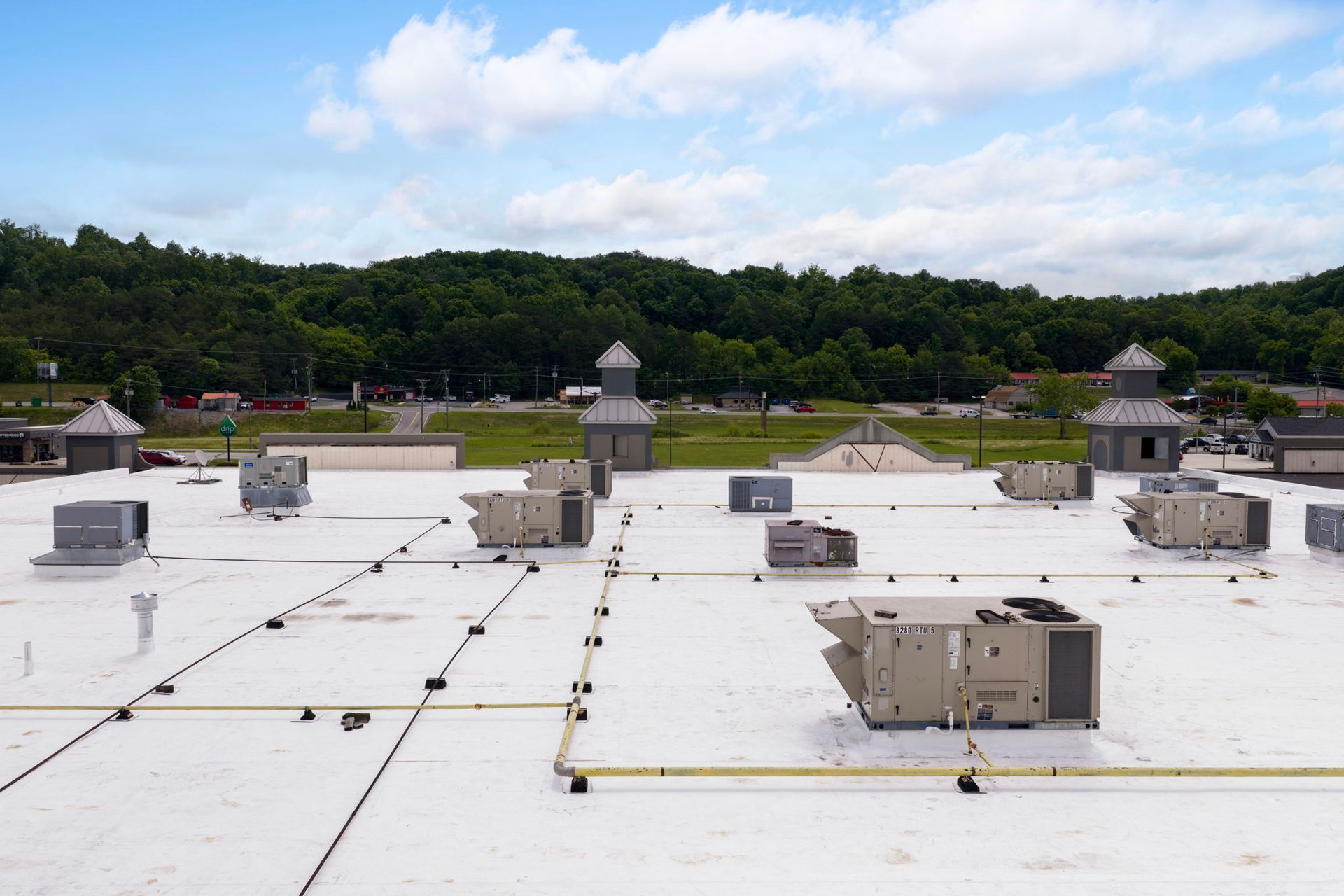 Flat white rooftop with vents and chimneys overlooking green trees under a blue sky with clouds.