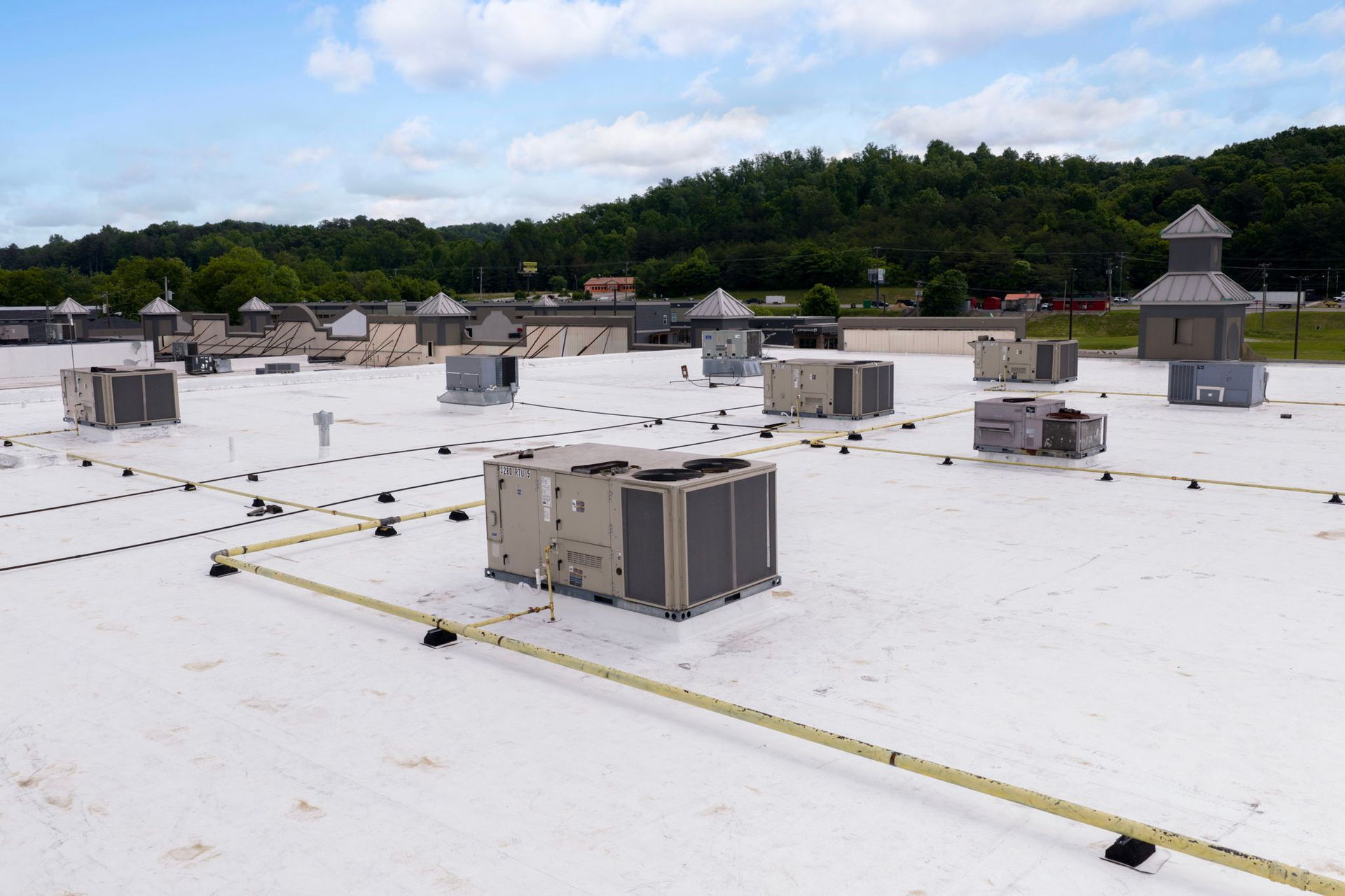 Flat white rooftop with several HVAC units under a cloudy sky, with trees and hills in the background