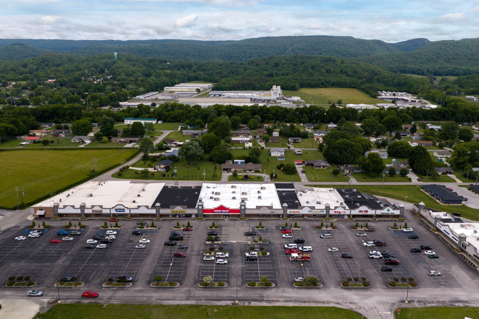 Aerial view of a shopping center with a large parking lot and surrounding green hills.