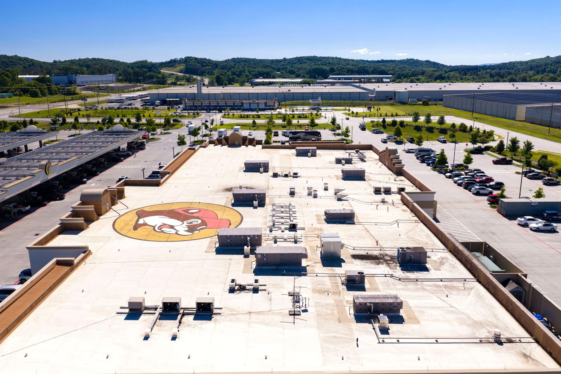 Aerial view of a flat rooftop with HVAC units and a large painted logo, surrounded by parking lots and trees