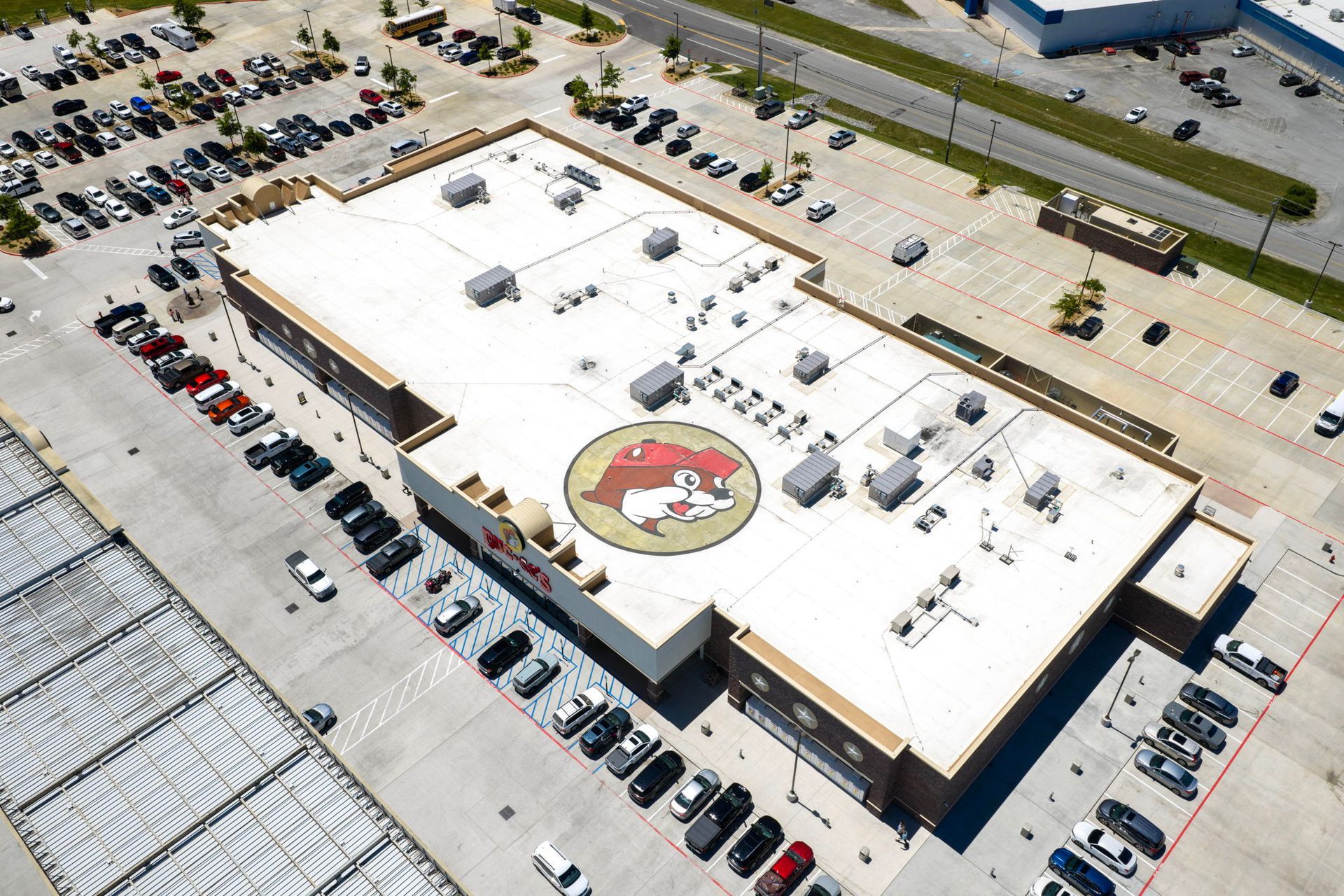 Aerial view of a large white-roofed building with a circular logo, surrounded by parked cars and roads.