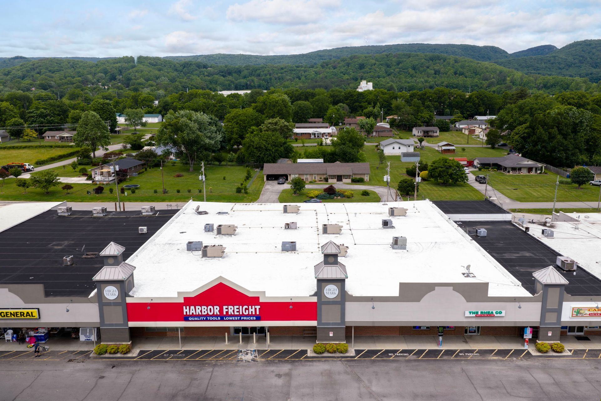 Aerial view of a strip mall with a white roof, red storefront sign, and parking lot in front of green hills.