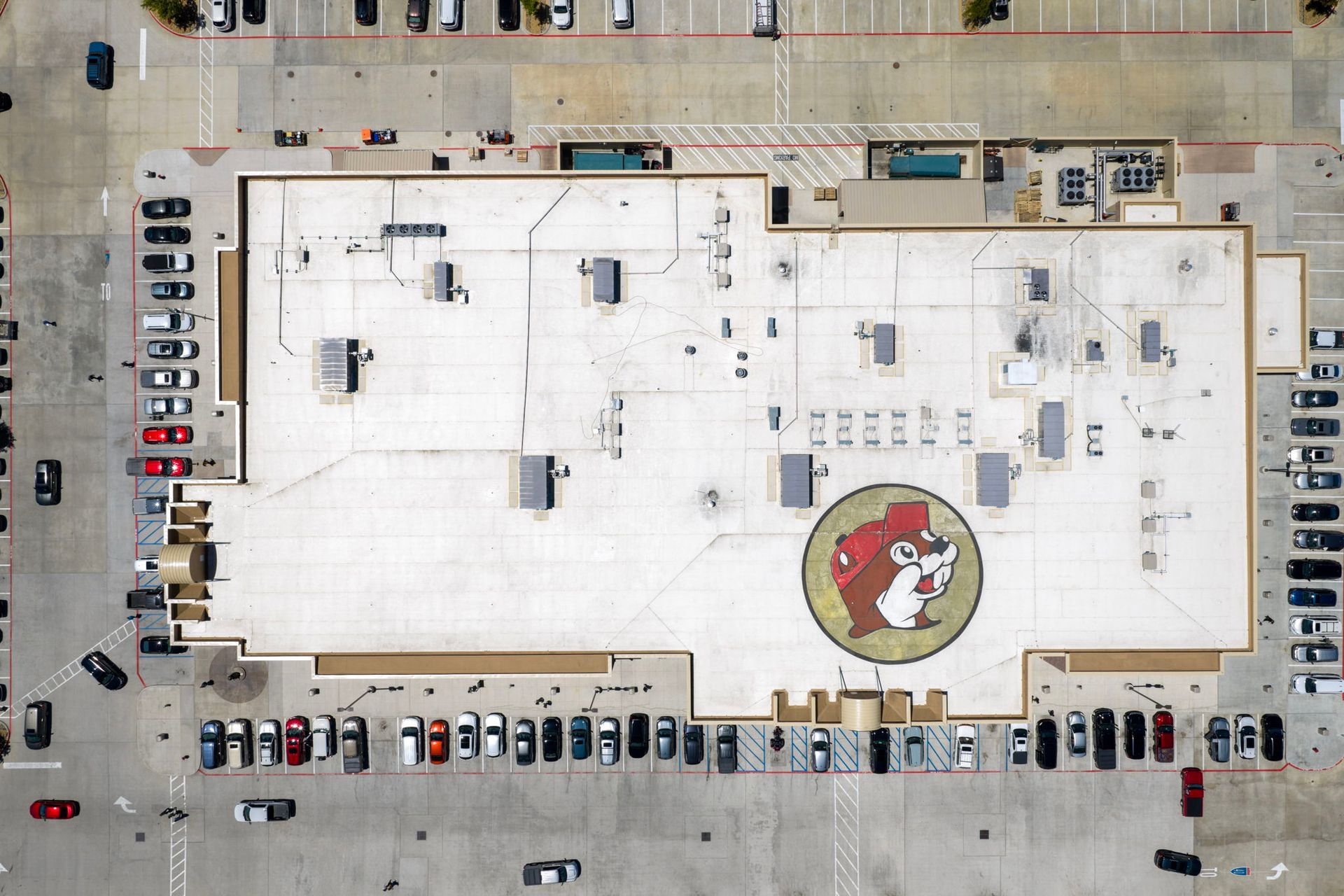 Aerial view of a large white-roofed building with parked cars around it and a circular logo near the center.