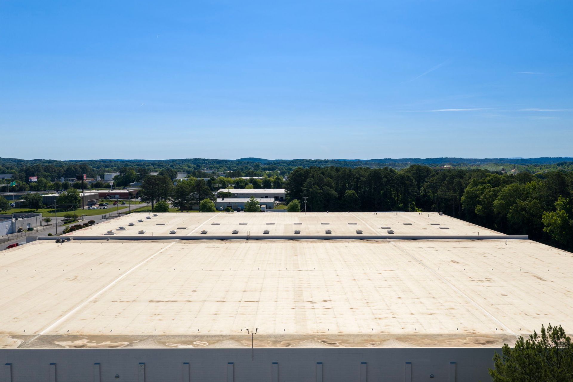 Aerial view of a large white warehouse roof beside trees under a clear blue sky