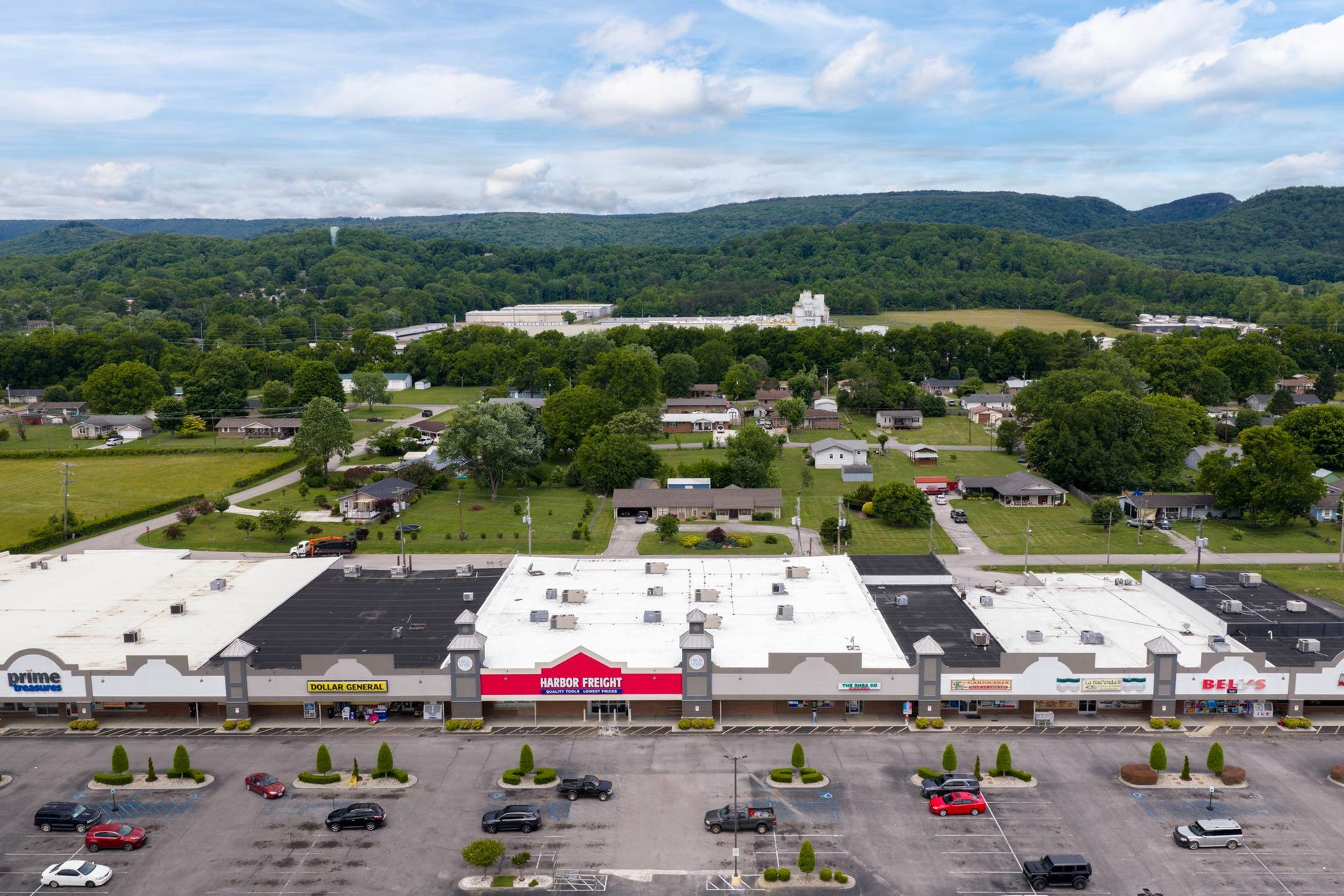Aerial view of a shopping center with a parking lot, surrounded by trees and hills under a cloudy sky