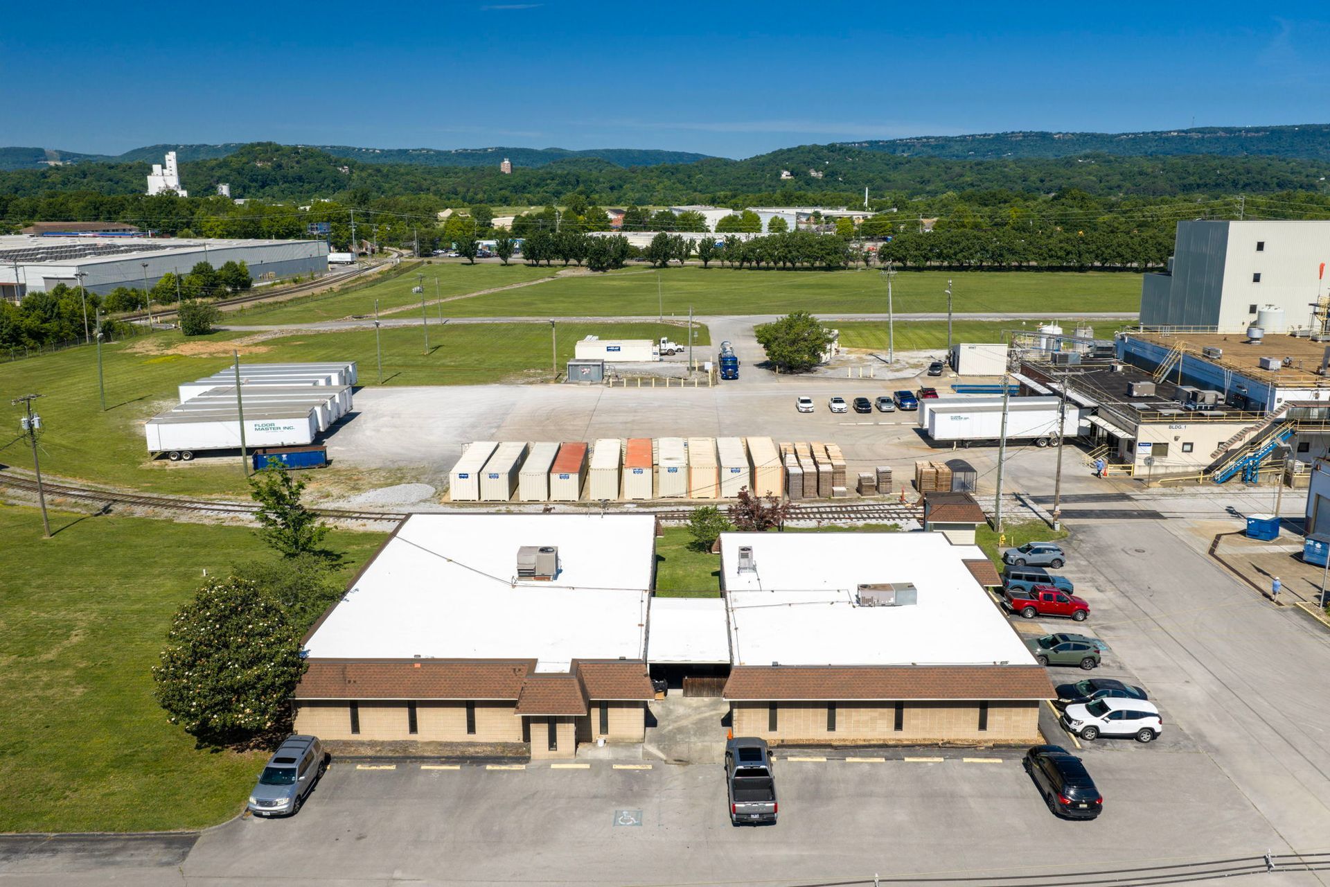 Aerial view of a small industrial yard with parked trucks, trailers, and warehouse buildings beside open fields.