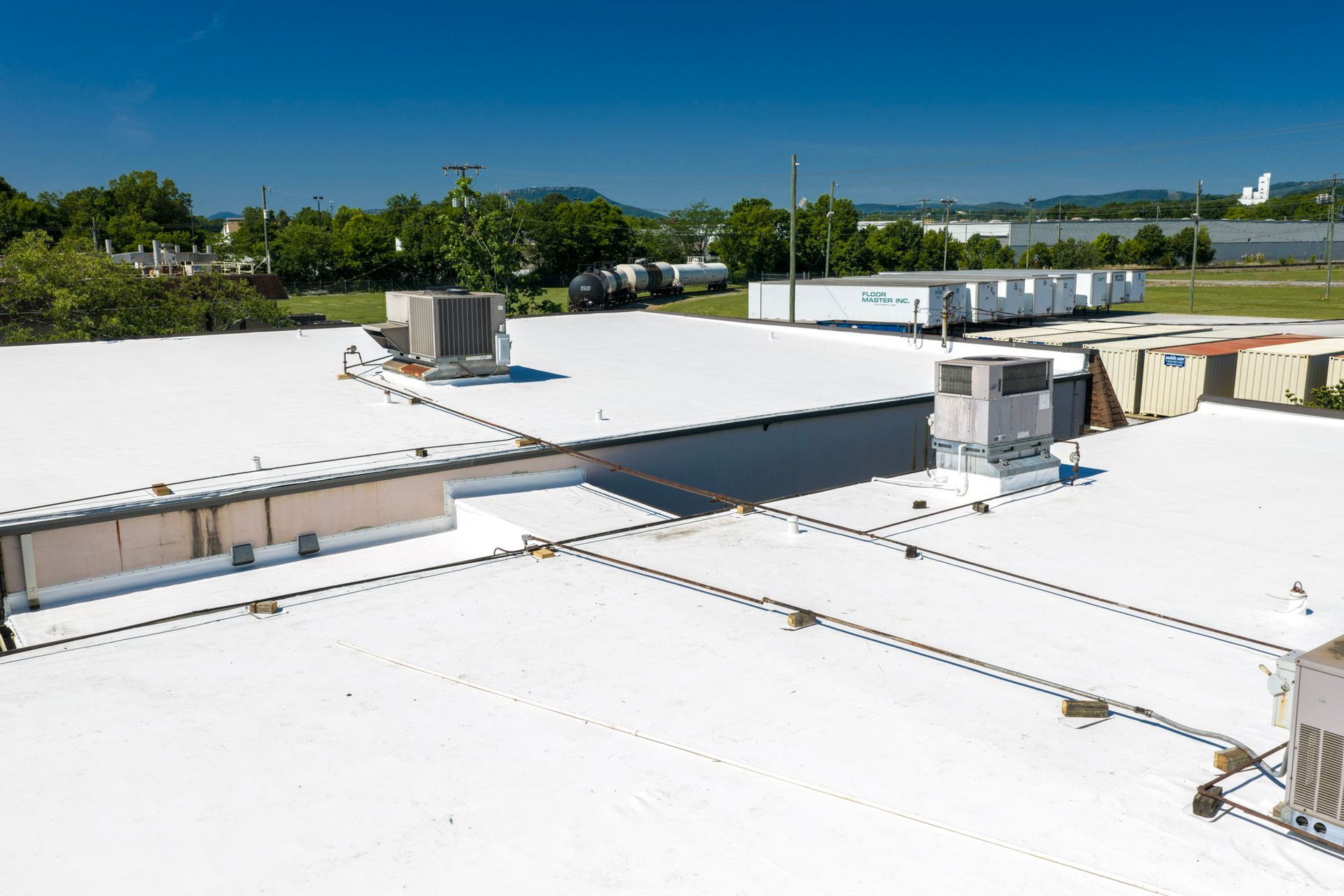 White flat rooftop with vents and HVAC units beside trees and water under a clear sky