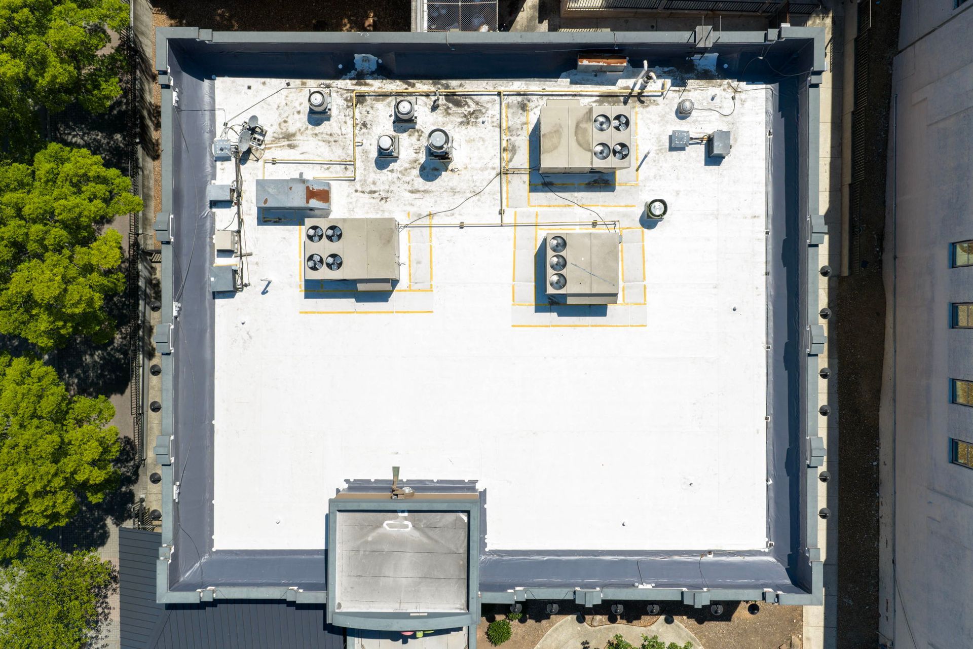 Aerial view of a flat white rooftop with HVAC units, vents, and surrounding trees and buildings.