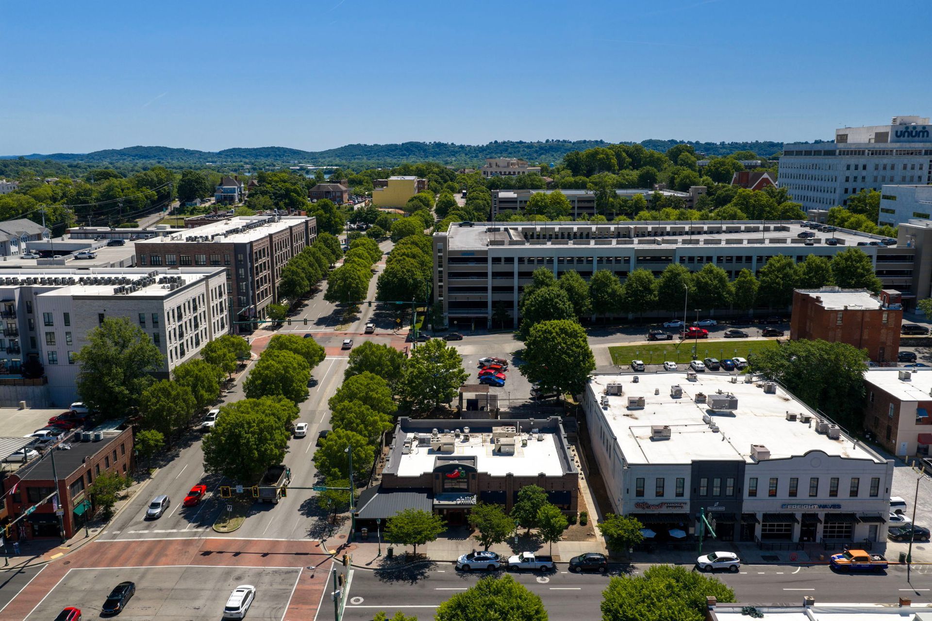 Aerial view of a downtown street lined with trees, low-rise buildings, and parked cars on a sunny day