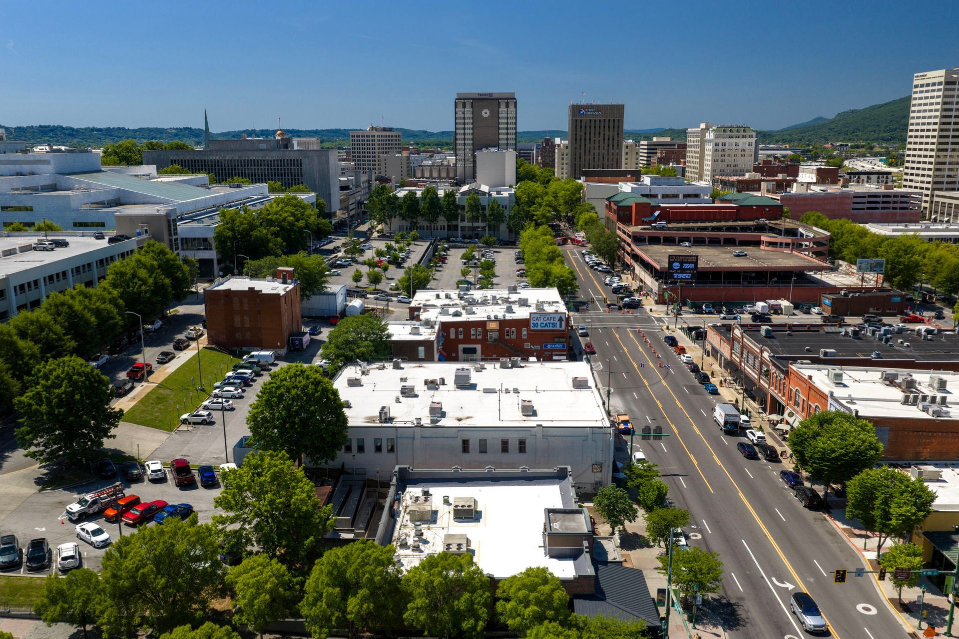Aerial view of a city street with buildings, trees, parked cars, and light traffic under a blue sky