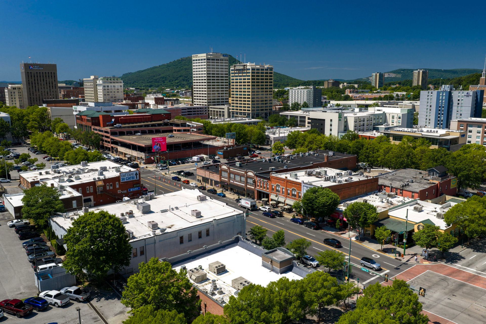 Aerial view of a city street with low-rise buildings, trees, and distant high-rises under a clear sky