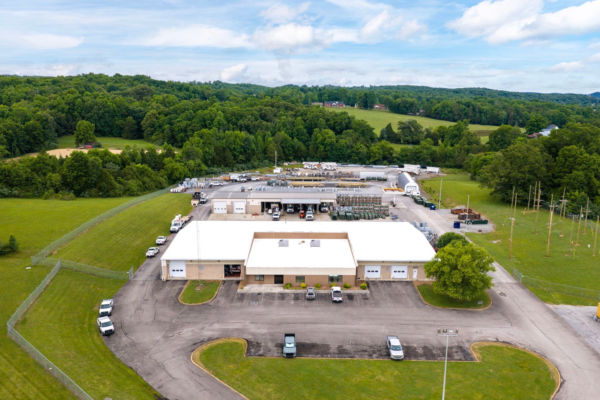 Aerial view of a white industrial building with parked cars, surrounded by trees and green fields.