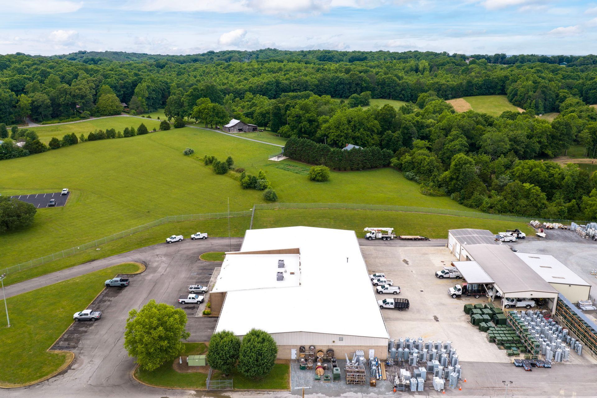 Aerial view of a white warehouse amid green fields and trees, with trucks and equipment in the lot
