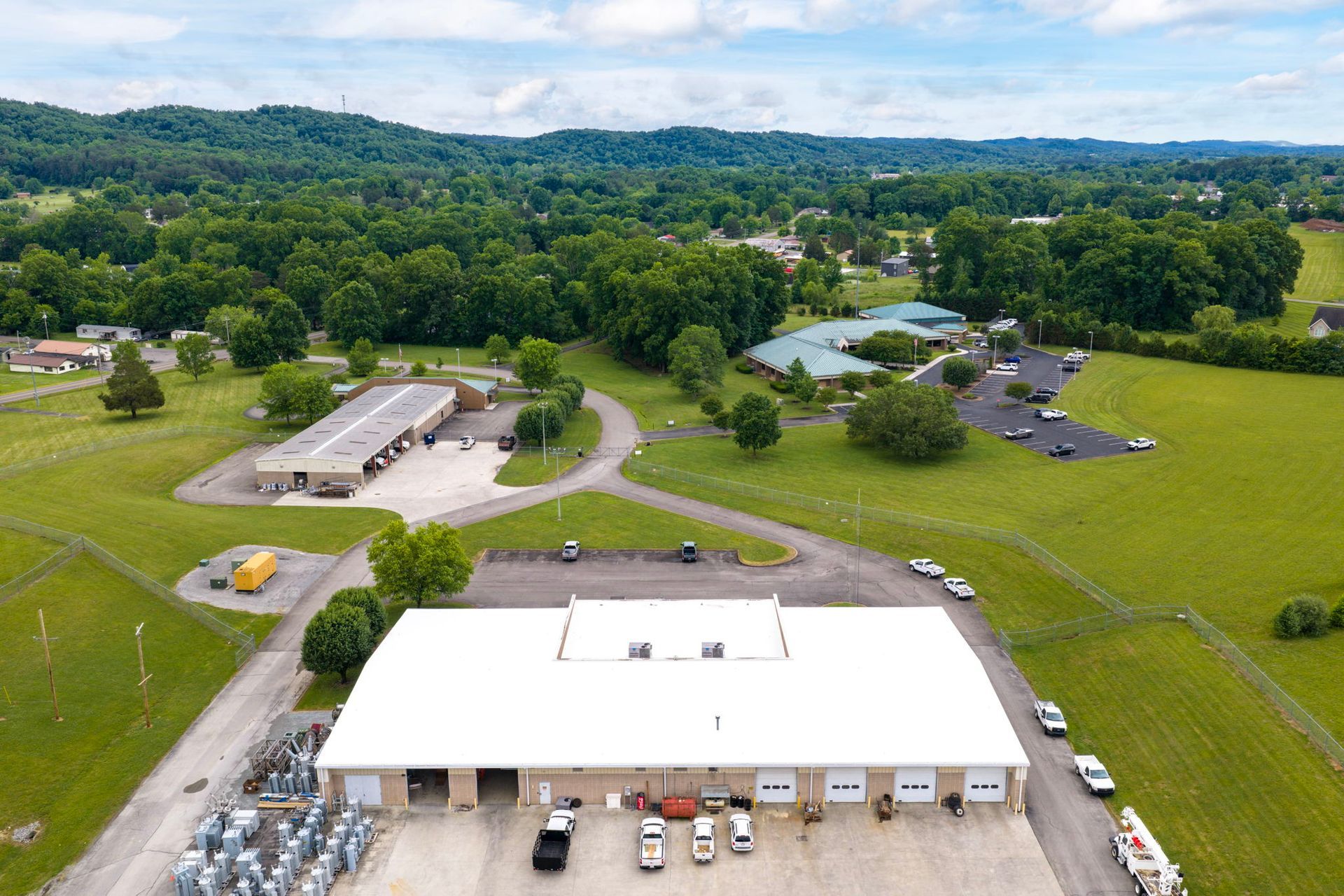 Aerial view of a warehouse complex with green fields, parked cars, and trees on a cloudy day