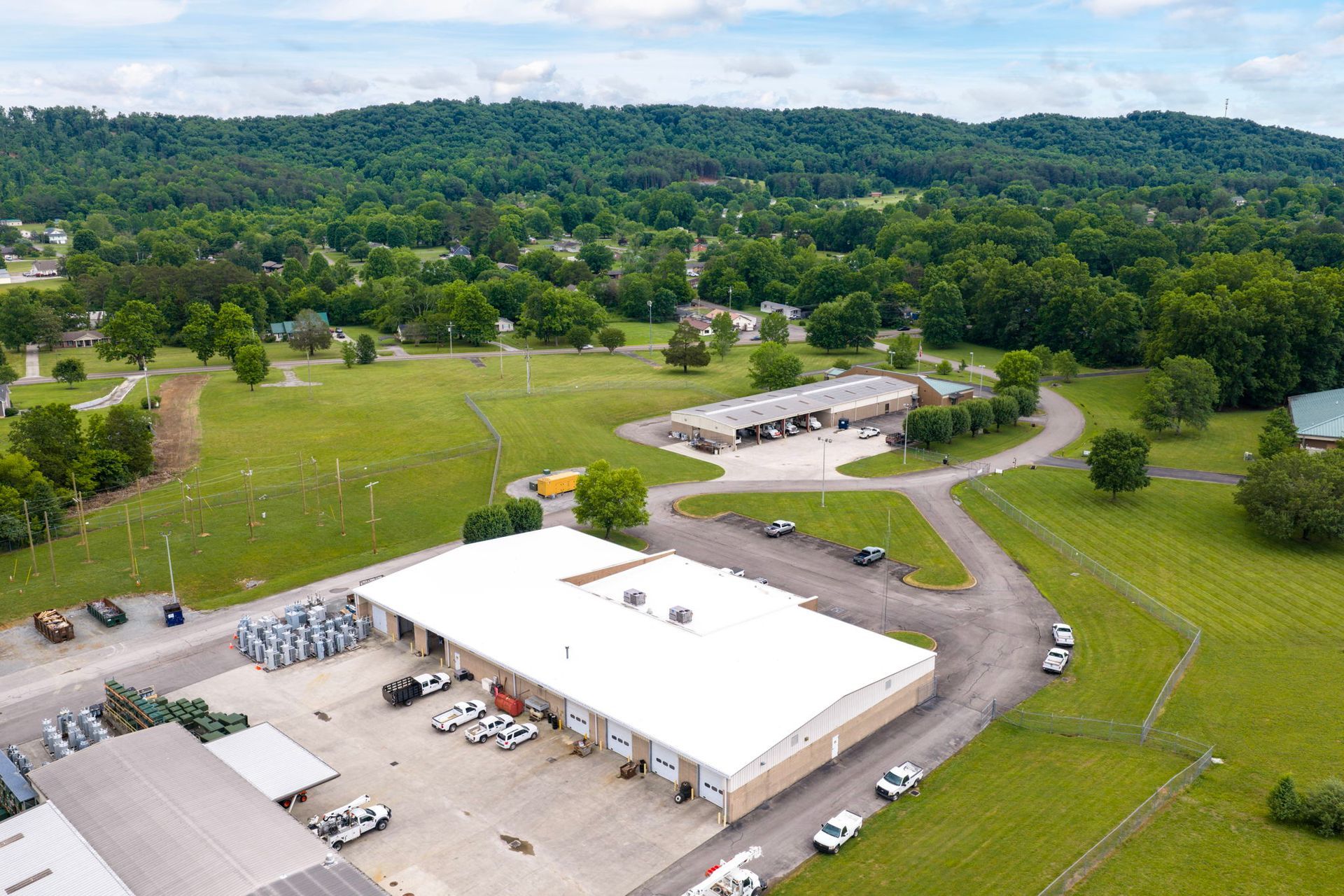 Aerial view of a white-roofed building, parked vehicles, and a green campus with trees and open fields