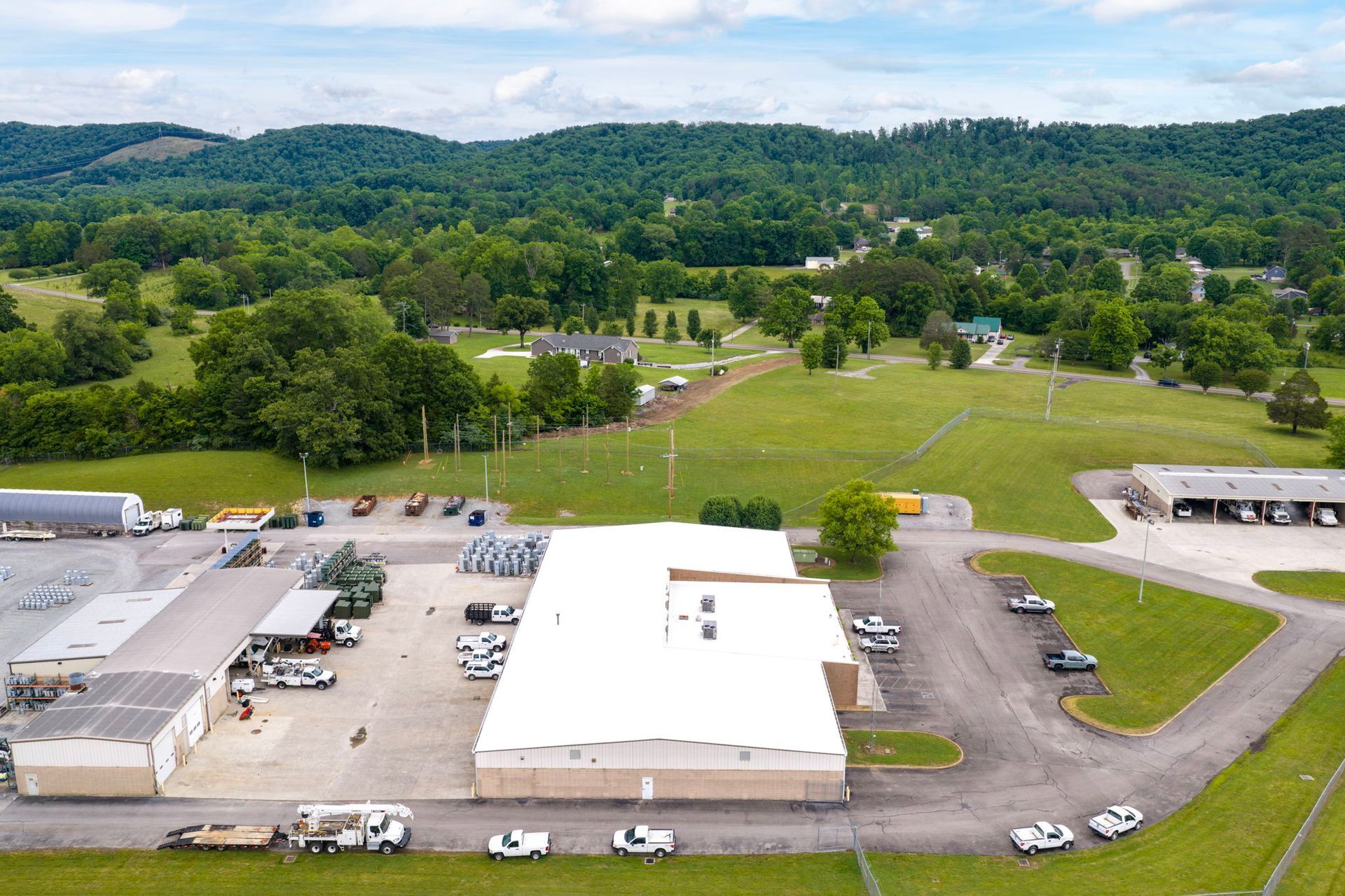 Aerial view of a rural campus with white buildings, parking lots, green fields, and wooded hills.