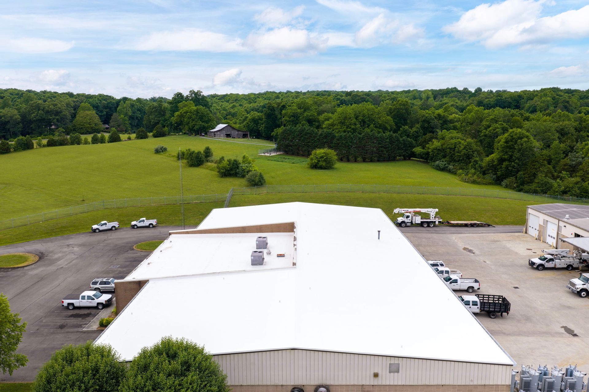 Aerial view of a white warehouse and parking lot beside green fields and trees under a blue sky.