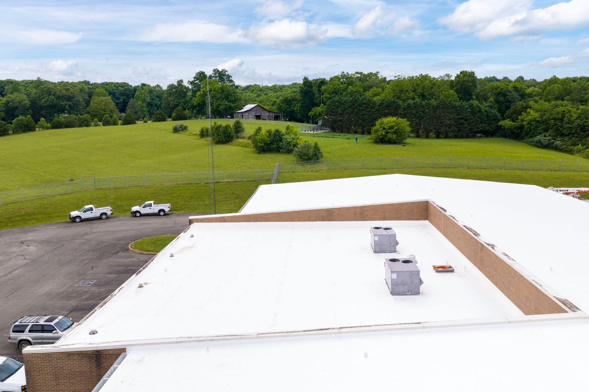 Rooftop with white membrane, HVAC units, and parking lot beside green fields and trees under a blue sky