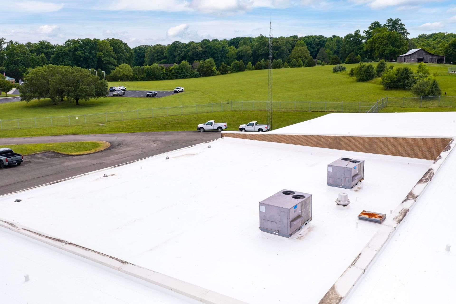 Flat white rooftop with two HVAC units, overlooking a green field and trees under a cloudy sky.
