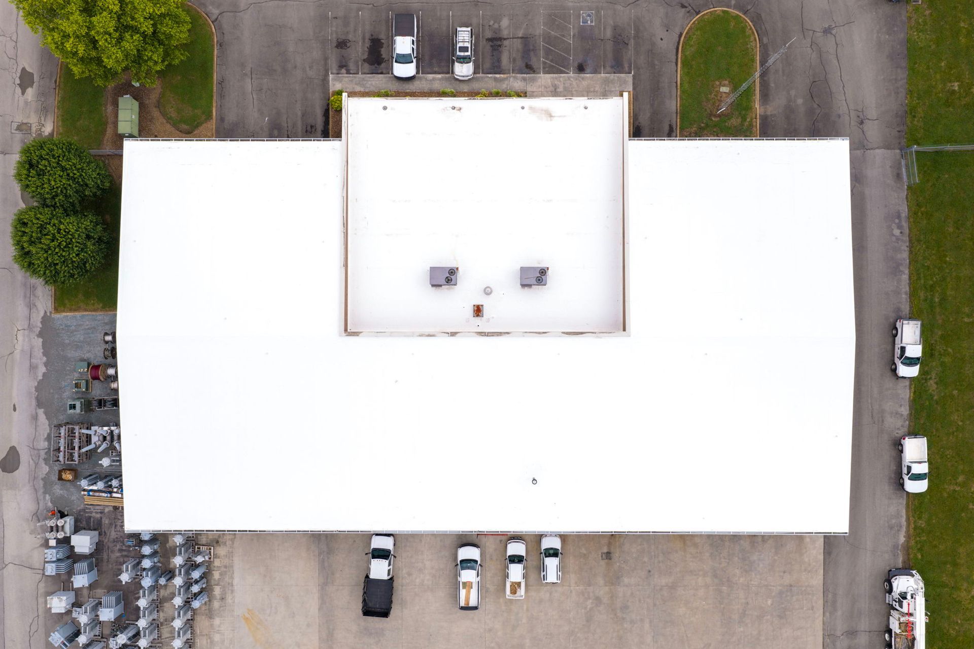 Aerial view of a large white-roofed building with parked cars around it and trees nearby