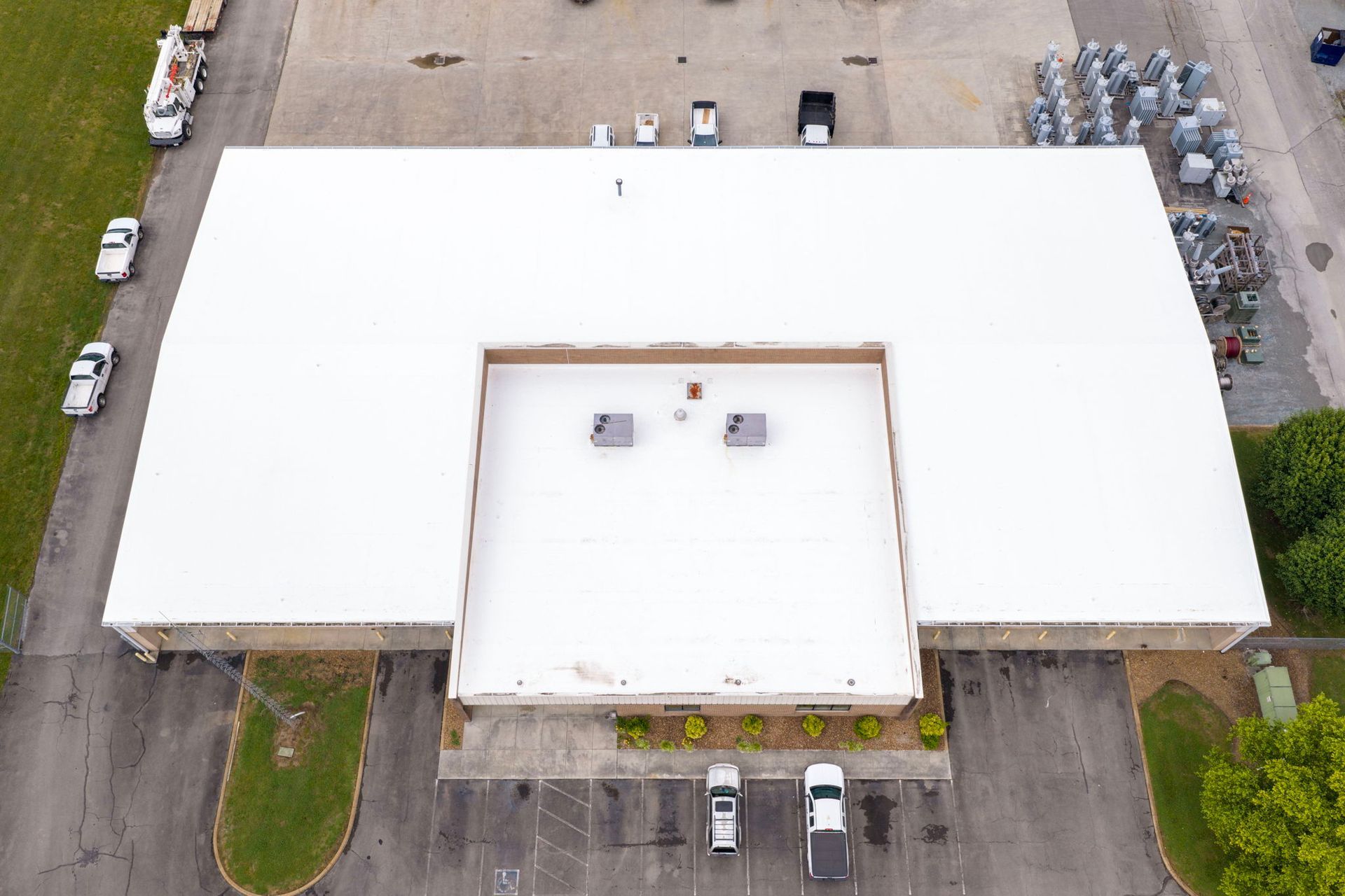 Aerial view of a large white-roofed industrial building with parking lots and trucks around it.