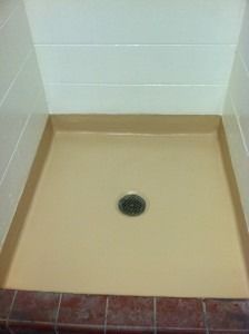 A square, beige shower pan with a central drain, framed by white tiled walls and a row of dark red floor tiles.