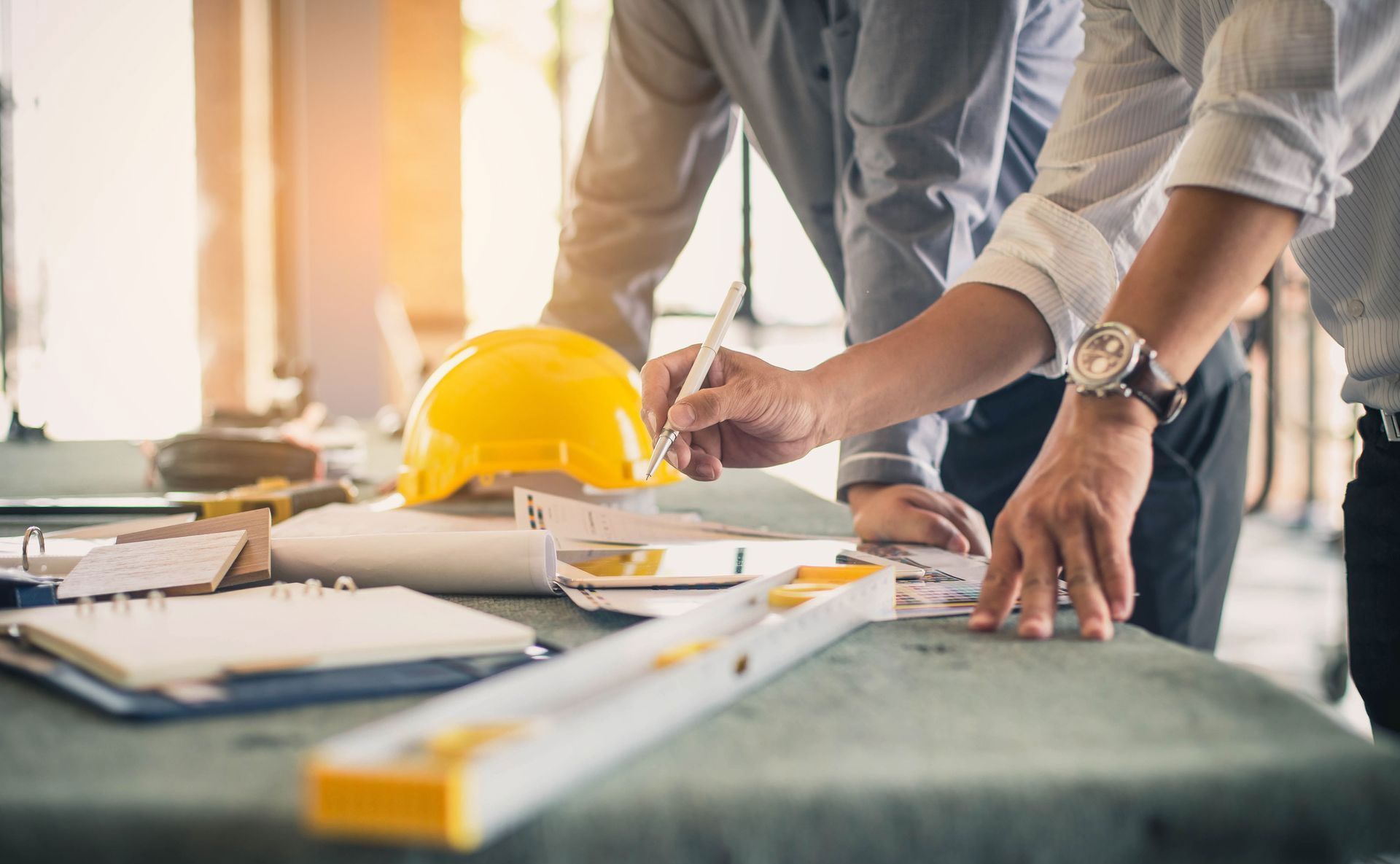 Two people in professional attire review blueprints on a table with a yellow hard hat and a level.