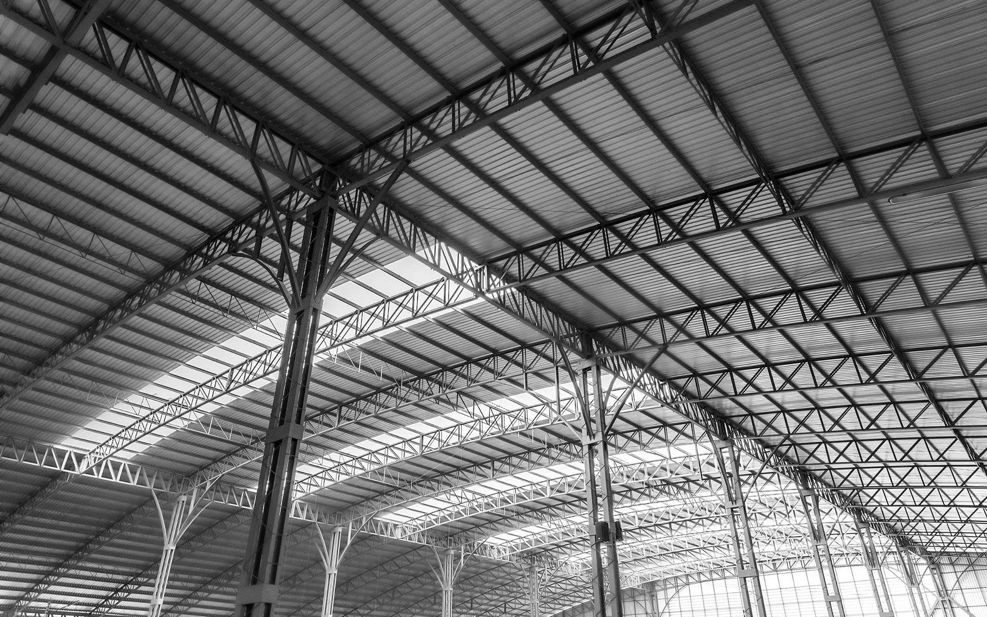 Low-angle, monochrome view of an industrial building's interior featuring an expansive metal truss and corrugated roof.