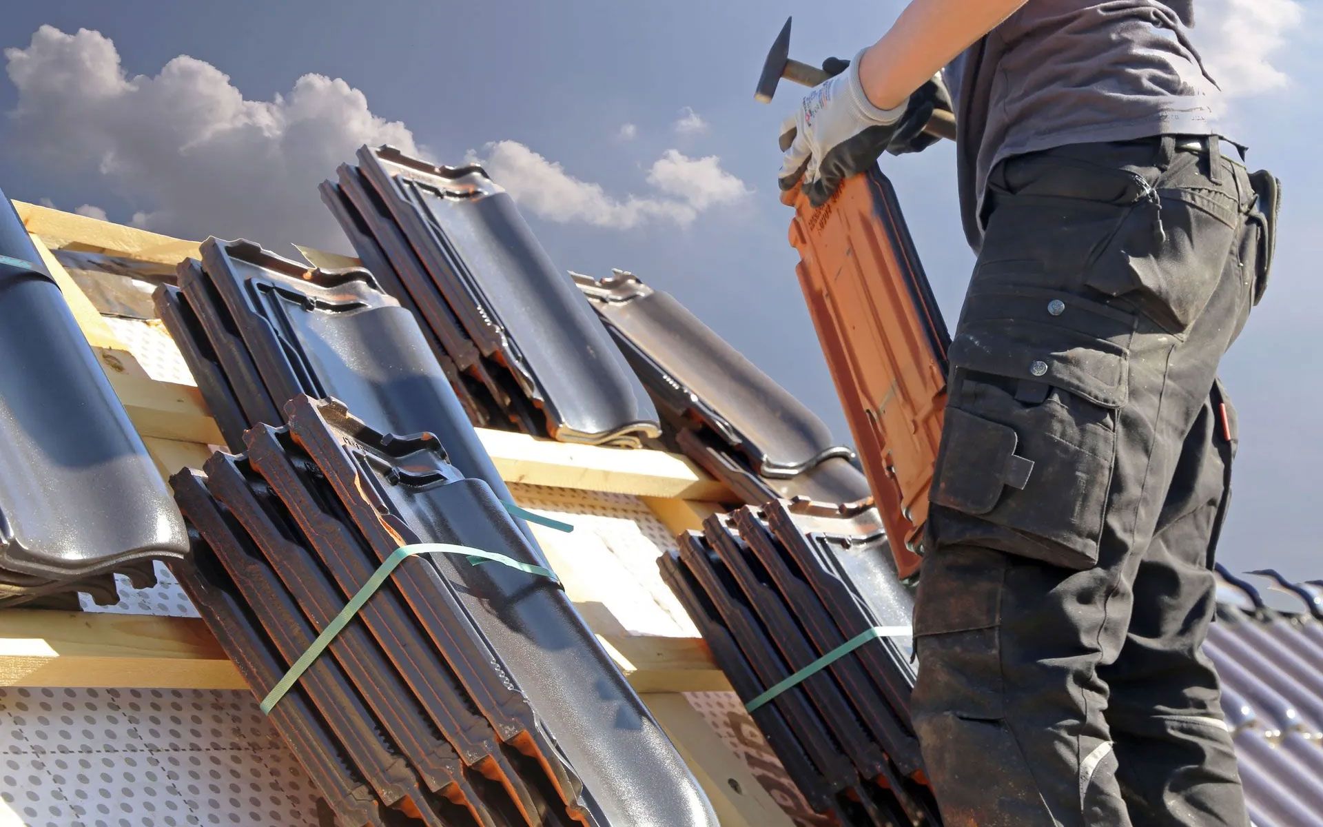 A roofer wearing gloves stacks dark, curved roof tiles on a wooden frame against a partly cloudy sky.