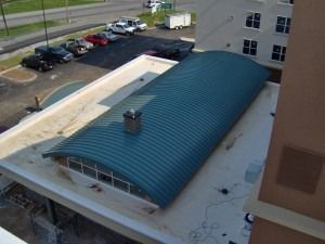 High-angle view of a building with a curved teal metal roof, a chimney, and a flat white roof section next to a parking lot.