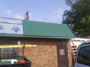 A worker in a white shirt stands on a brick building's roof installing green metal panels next to uncovered roofing paper.