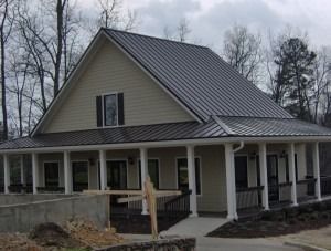 A beige house with a dark metal roof, wrap-around porch with white columns, and a partially finished concrete patio area.